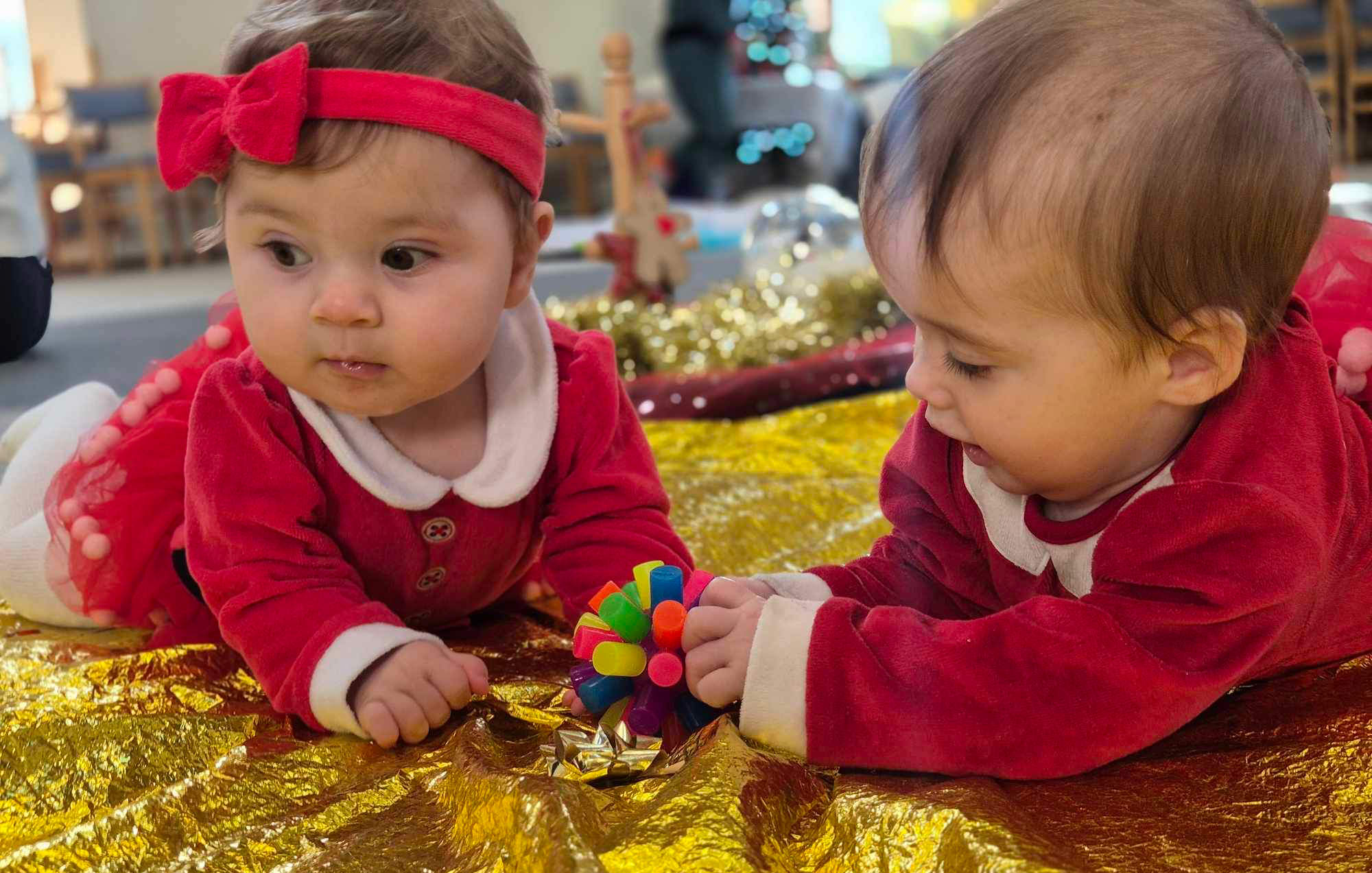 Babies in festive red outfits playing with a colourful toy on a gold play mat